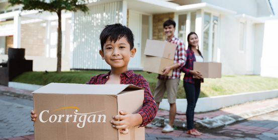 A boy holds a box printed with “Corrigan,” smiling at the camera. His parents carry other boxes behind him, outside a new house.